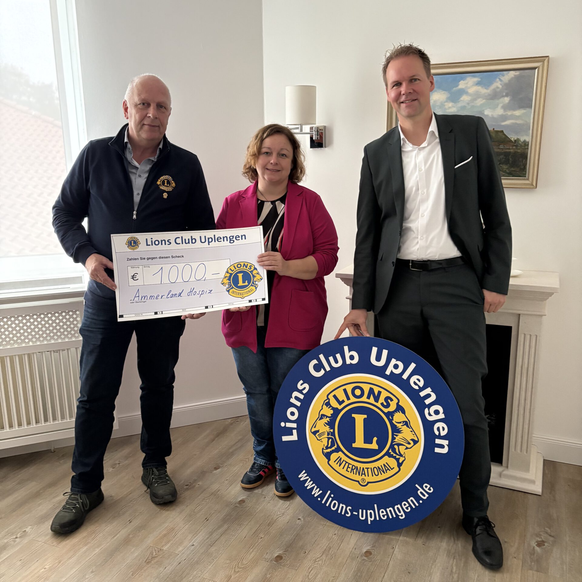 Spendenübergabe des Lions Club Uplengen an das Ammerland-Hospiz: drei Personen mit einem symbolischen Scheck über 1.000 Euro und dem Lions-Club-Schild.
